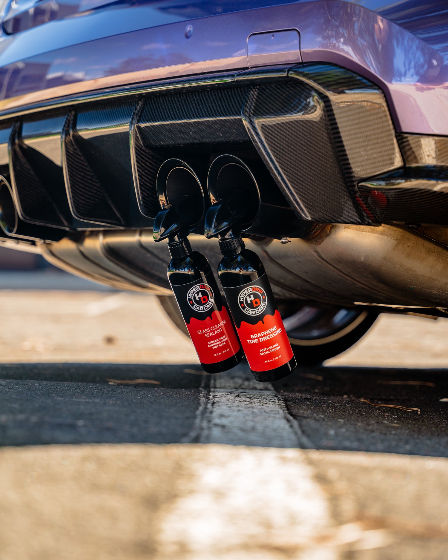 HyperDrip Glass Cleaner and Graphene Tire Dressing bottles hanging from the carbon-fiber rear diffuser of a purple sports car.